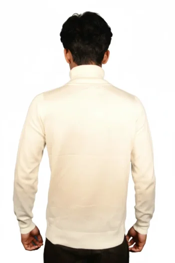 A man stands facing away from the camera, wearing a long-sleeved, light-colored turtleneck sweater and dark pants against a plain white background.